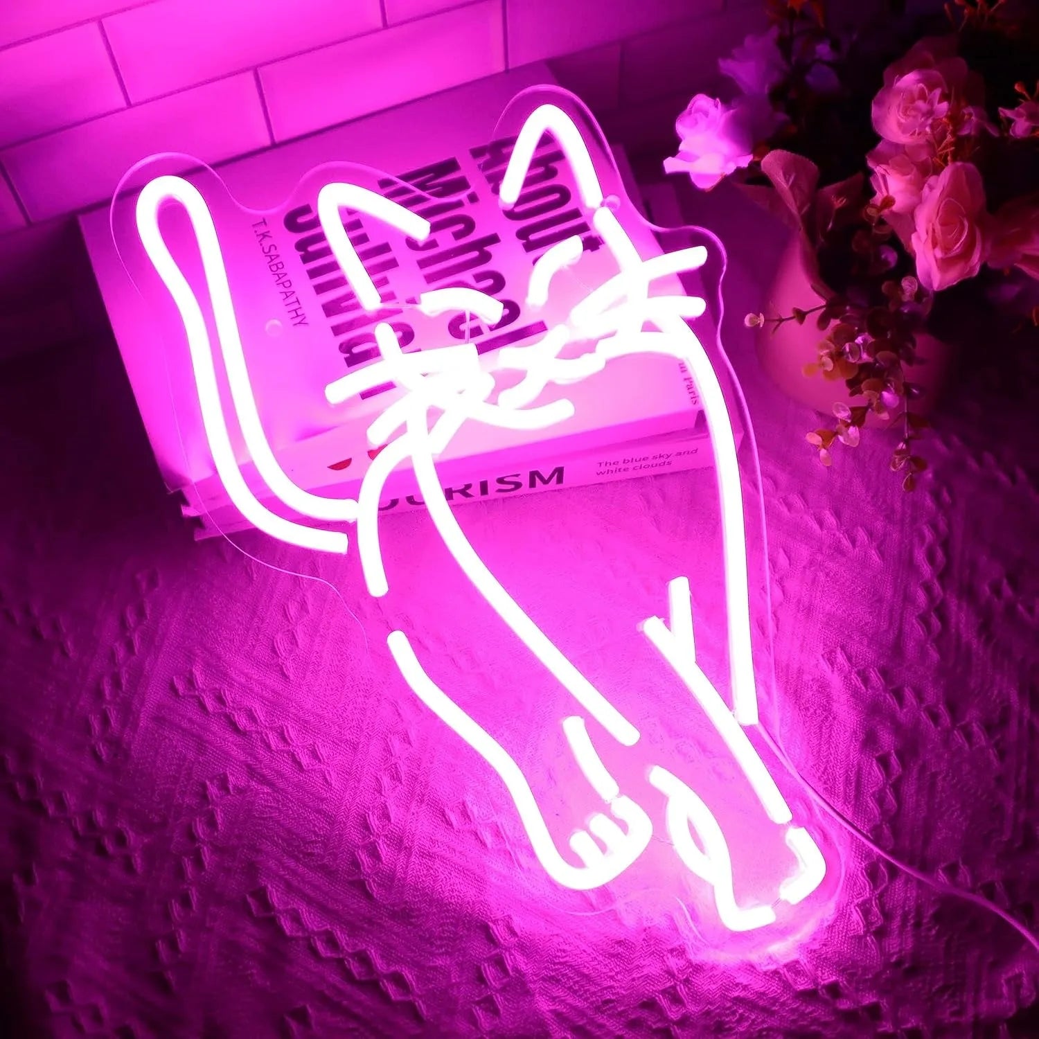 A vibrant pink LED neon sign of a cat mid-stride, displayed on a table with books and flowers.